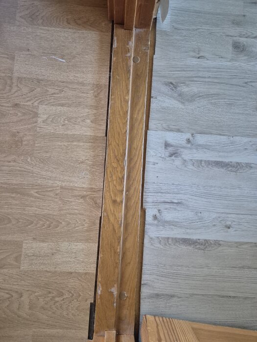 Två olika golv med en trätröskel emellan, där tröskeln är skadad och ojämn vid dörrkarmen.