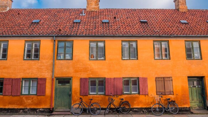 Gul-orange fasad med gröna dörrar, röda fönsterluckor och tegelpannetak. Tre cyklar står lutade mot väggen.