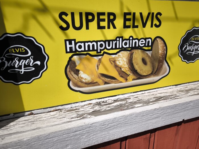 Reklamskylt för Super Elvis hamburgare med ananas och ost, från Elvis Burger, på en gul bakgrund.