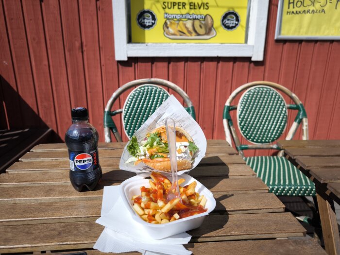 Elvisburgare med pommes och Pepsi Max på ett träbord utomhus, skylt med "Super Elvis" i bakgrunden.