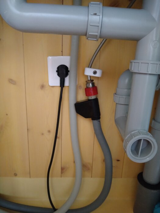 Diskmaskinens slangar och aquastop under diskbänken med synliga avloppsrör och vägguttag i en sommarstuga.