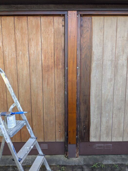 Två garageportar med färgskiftningar, vänster port tvättad, höger port slipad. Planka mellan portarna oljad. Stege med färgburk syns.