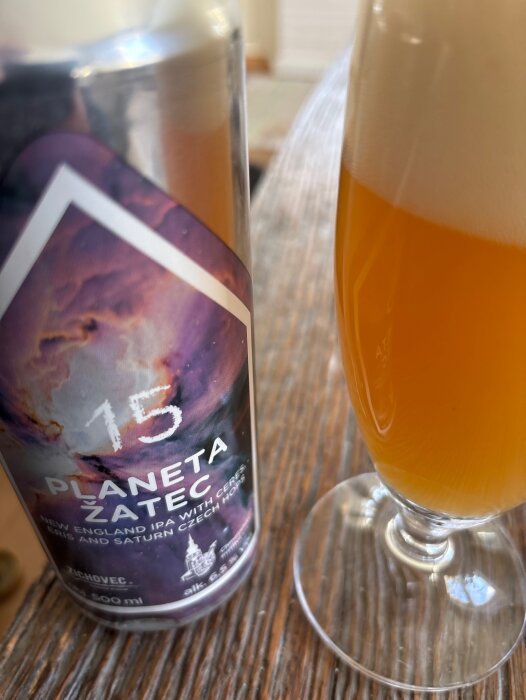 Ölburk med etiketten "Planeta Zatec" och ett glas med skummande orangefärgad IPA står på ett bord inomhus.