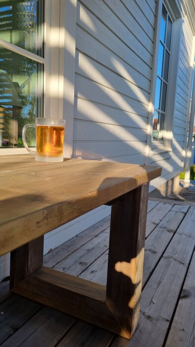 En kall öl i glas på ett träbord, solen skiner, och vita träpaneler i bakgrunden.