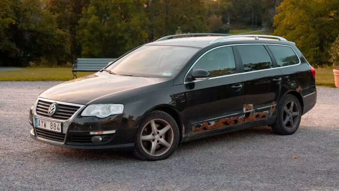 Svart station wagon med rostskador längs sidan, parkerad på en grusväg.