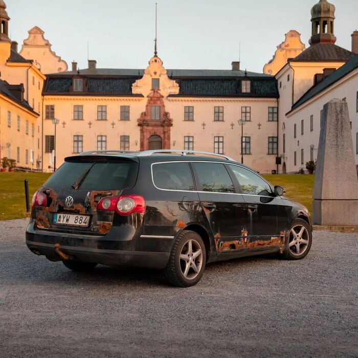 Svart Volkswagen med rostskador parkerad framför ett stort vitkalkat husliknande byggnad.