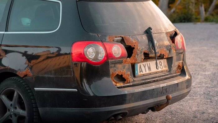 Bakdelen av en bil med omfattande rostskador runt baklyktor och registreringsskylt, mot en grusväg och träd i bakgrunden.