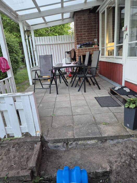 Uteplats med stolar och bord på stenplattor intill ett hus med rött träfasad och vita knutar.