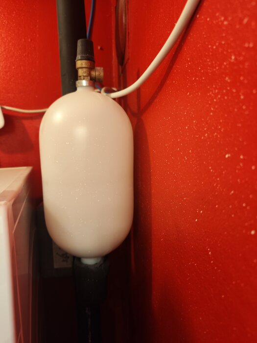 Expansionskärl under jordvärmeslinga med rött vägg i bakgrunden, utrustat med rör och anslutningar.