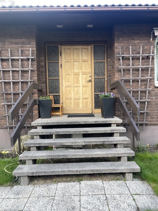 Trappuppgång till ett hus med träfasad och entrédörr. Två blomkrukor med blommor står vid sidan om dörren.