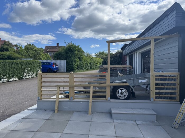 Träställning byggd på en garageuppfart, med en släpkärra bakom. En blå bil och grönska i bakgrunden under en molnig himmel.