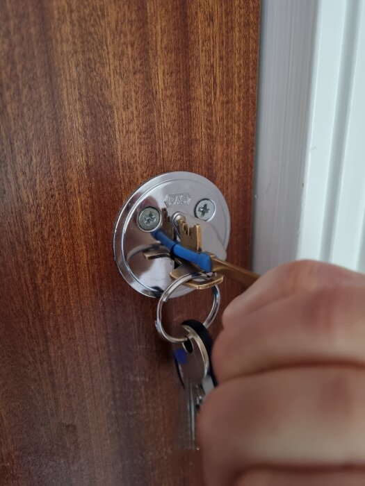 Nyckel insatt i låscylinder på en brun dörr, fasthållen av en hand, med flera nyckelringar fästa.