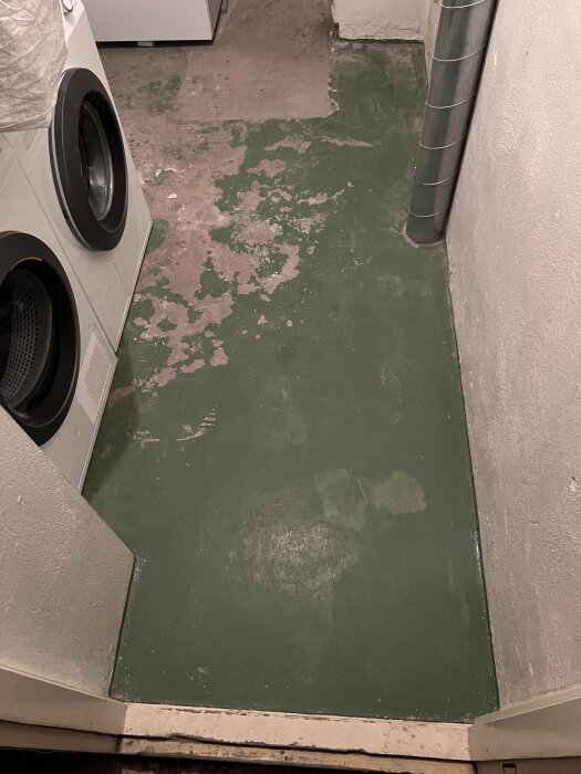 Källare med grön målad betonggolv, flagnande färg, tvättmaskin och ventilationsrör synliga.