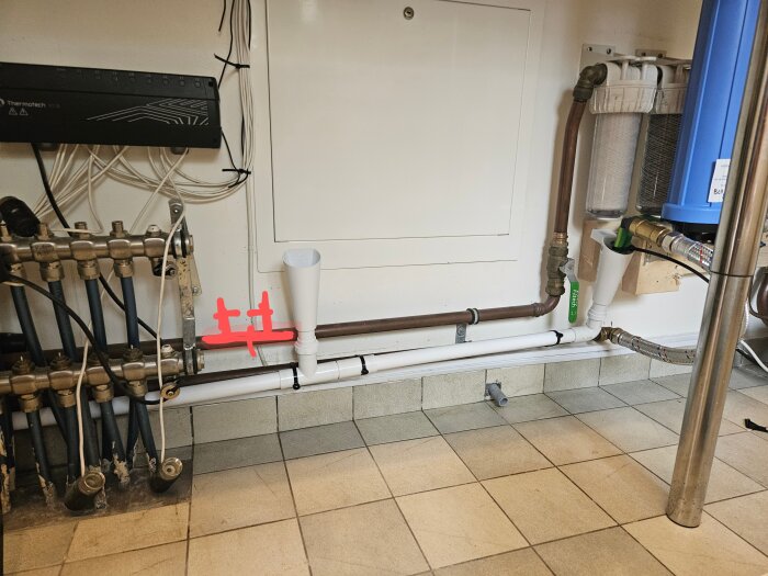 Rörsystem med kopplingar nära golvvärmefördelare, visar luftblandare och filter installerade på kopparrör samt en grön ventilmärkning.