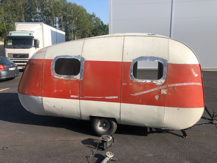 Gammal husvagn under renovering, med avskalad färg och arbetade fönsteröppningar; verktyg på marken, omgiven av andra fordon.