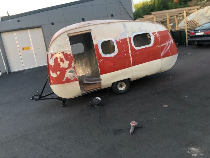 Renovering av rundad, röd-vitt sliten vagn på asfaltsplan, med öppning vid dörren och redskap runtom.