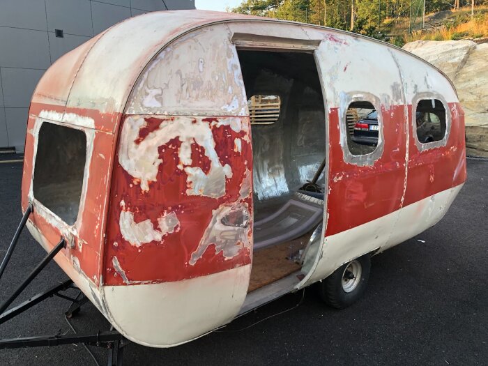 Sliten husvagn under renovering, delvis bortslipad färg och spackel på röda och vita ytor, tre fönsteröppningar, ett öppet hål i dörrpositionen.