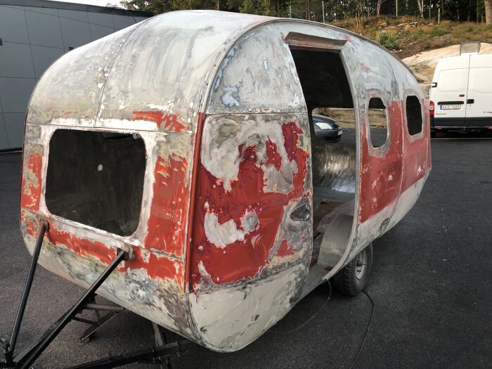 Renovering av husvagn med avskalad färg och spackling på karossen, utskuret runt fönster och dörröppning, belägen på asfalterad yta.