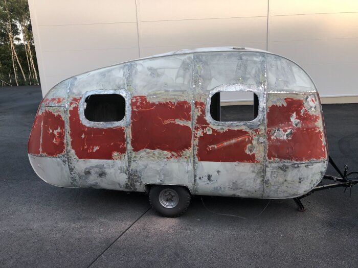 En husvagn under renovering med avskavd röd färg och öppna fönster, delvist slipad för att bli metallren, stående på en asfaltsyta.