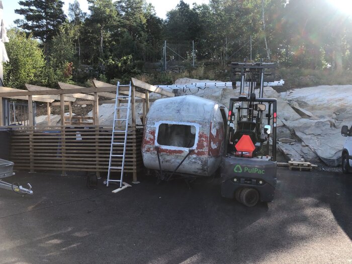 Trailer på gårdsplan under renovering med borttagning av gammal färg, omgiven av byggställning och gaffeltruck med solbelysta träd i bakgrunden.