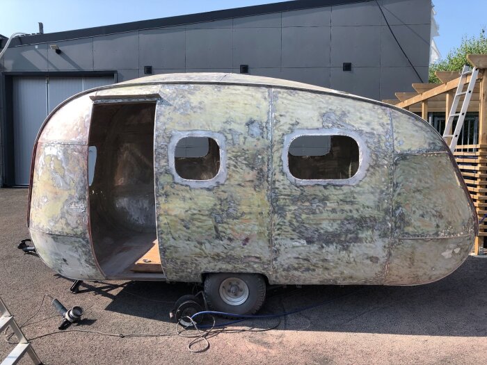 Renovering av husvagn i metall, med slipad utsida och öppna fönsteröppningar, står på en asfaltbelagd yta bredvid byggnader.