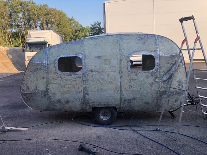 Renovering av husvagn pågår med slipad aluminiumyta, två fönster och byggställning. Vinkelkap och stege syns bredvid i solig miljö.