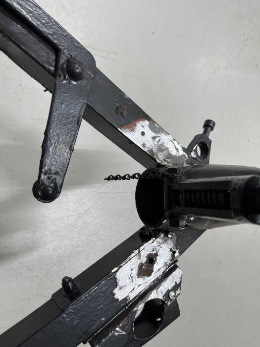 Svart metallstruktur med tecken på rost och färgbortfall, del av underrede på en vagn med synliga svetsningar och kedjor.