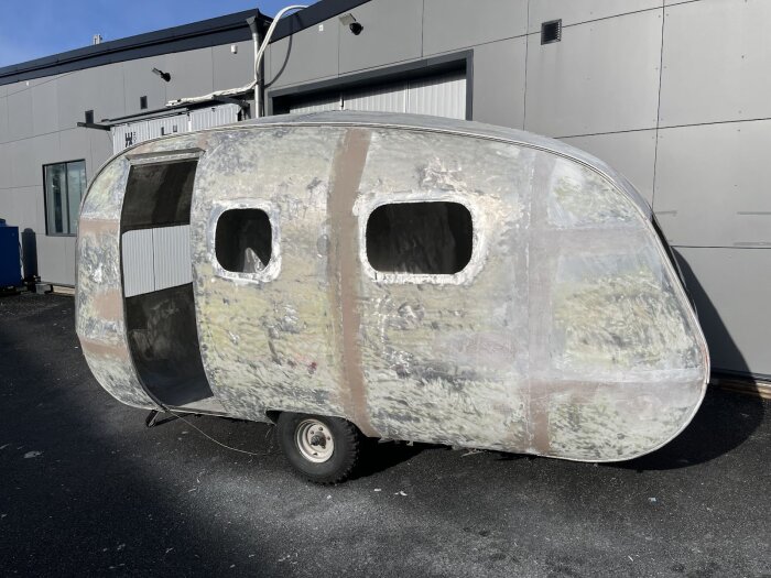 En renoverad husvagn med slipad och spacklad utsida, placerad framför ett grått industribyggnad. Fönster och öppningar synliga, inga dörrar monterade.
