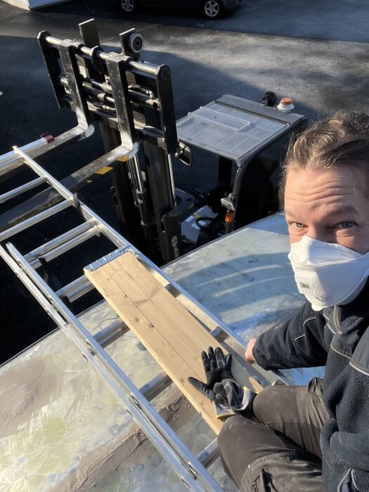 Person med ansiktsmask sitter på en provisorisk arbetsställning av plankor och metallstegar, stödd av en truck, under arbete på vagntak.