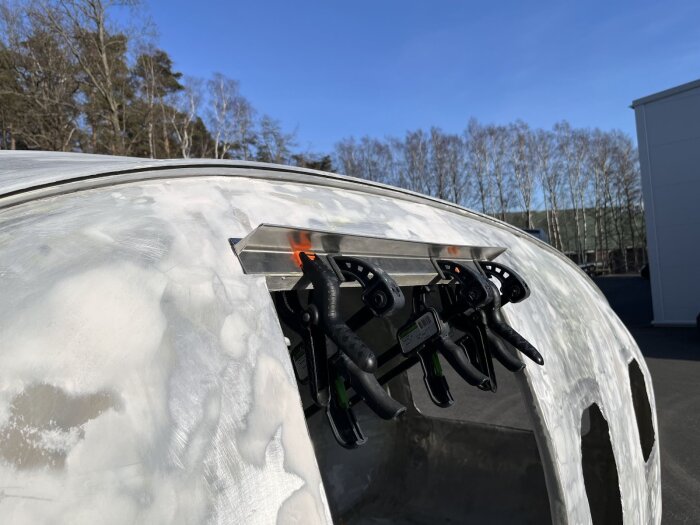 Slipad vagn med ny aluminiumbåge över dörren, säkrad med tvingar, under en klar himmel. Renoveringsarbete av utsidan pågår.