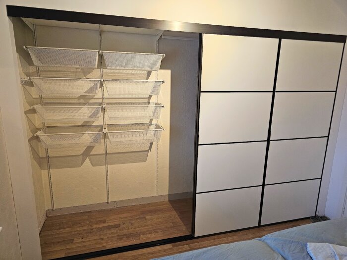 Väggmonterad förvaring med nio vita nätkorgar och skjutdörrar i vitt med svarta ramar, installerade i ett rum med trägolv.