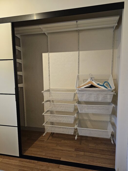 Vit garderobsinredning med flera nätkorgar och klädgalgar, monterad på dubbla vertikala väggskenor inne i en öppning med skjutdörrar.