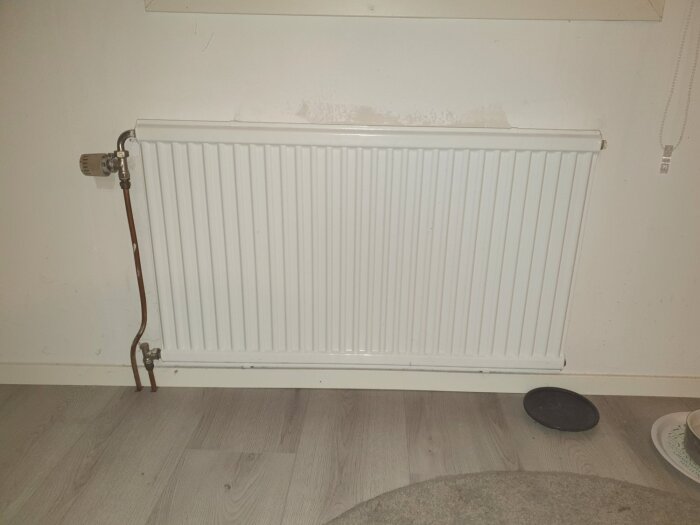 En vit vattenradiator installerad mot en ljus vägg, med kopparrör anslutna på vänster sida. Radiatorn ska ersättas med en större modell.