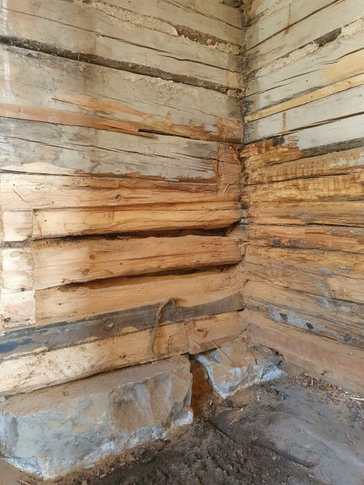 Skadat timmer i hörn av hus, delvis borttagna lager, synliga stenar i botten. Renovering pågår efter angrepp av myror eller vatten.