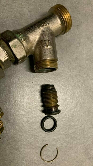 Returventil i närbild med separerade delar, inklusive gängade kopplingar och en O-ring, på ett grått underlag.