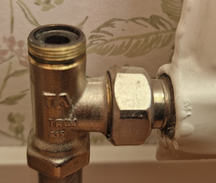 Returventil ansluten till en vit radiator med mönstrad tapet i bakgrunden.