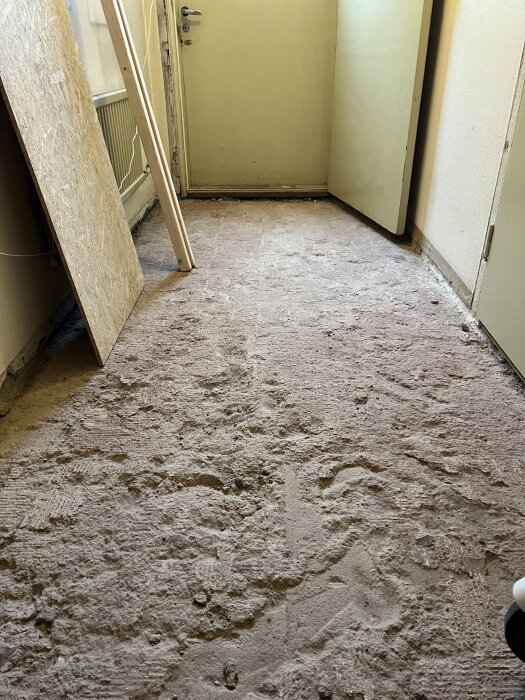 Riven klinker i korridor på bottenvåning, exponerat grovt golv från 60-tal med byggdamm och byggmaterial lutande mot väggen.