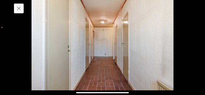 Gammal korridor med avskalad golvyta där klinker har tagits bort, vita väggar, flera dörrar och taklampa synliga.
