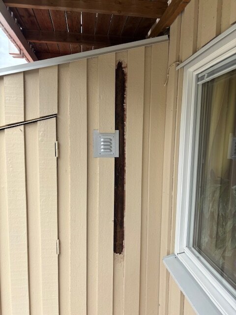 Exponerad plywoodyta med bortskrapad färg och lockpanel, synlig under ett balkongtak; ventillation med svart mögel på träytan.