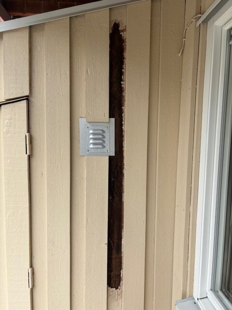 Exponerad plywoodyta med bortskrapad färg och svartmögel synligt på väggen under en balkong med lockpanel, bredvid en ventil.