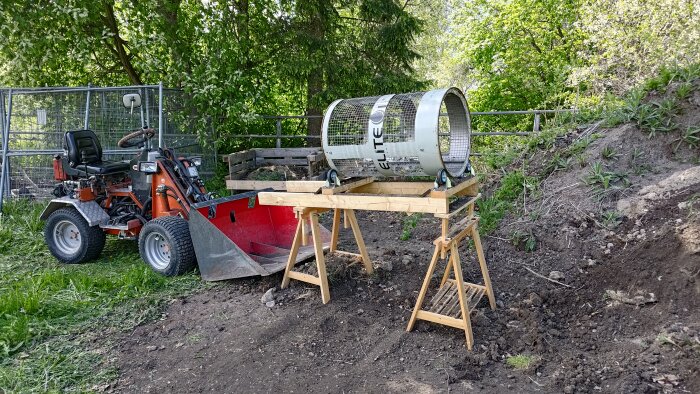Manuell trumma monterad på stativ bredvid traktor och jordhög, omgiven av grönska.