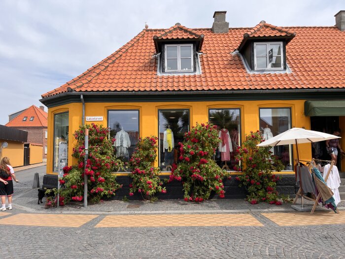 Gult hus med röda rosor och skyltfönster vid Laksetorvet. Klädhängare och parasoll utanför, personer i närheten.