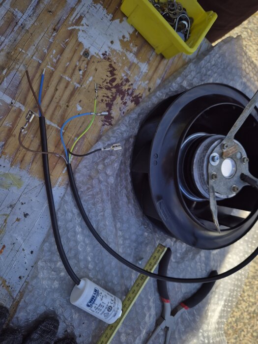 Nya fläkten på träbord med synliga kablar och kondensator, gula plastlåda med metalldelar, mätband och tänger ligger bredvid.