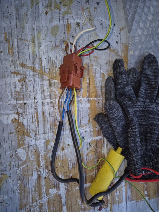 Kablar kopplade i en brun kopplingskontakt på sliten träyta, omgivna av arbetshandskar och en gul komponent, eventuellt en kondensator.