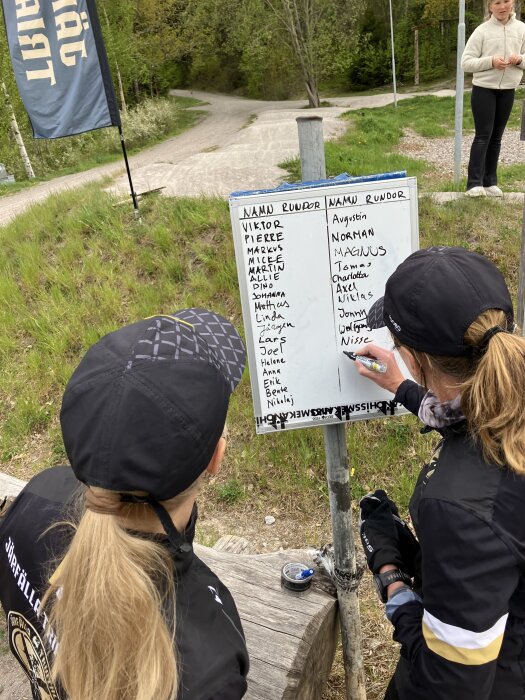 Personer vid en skylt utomhus under en tävling, skriver och tittar på resultatlista i blåsig och regnig miljö.