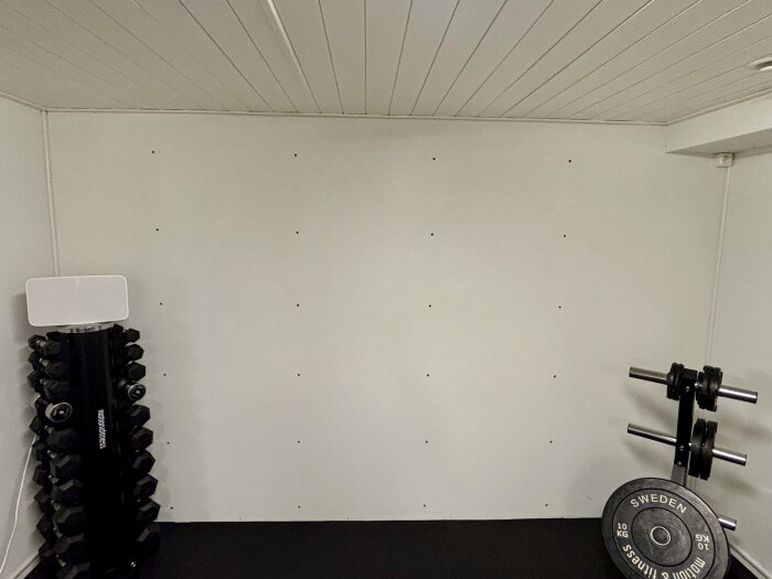 Vit vägg med hål från tidigare hyllsystem i källargym, omgiven av hantlar och viktskivor.