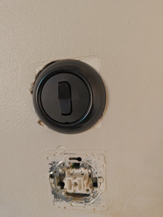 Rund strömbrytare av märket Renova installerad på ojämnt vägghål, med en öppen dosa under, där fyrkantig passform tidigare funnits.