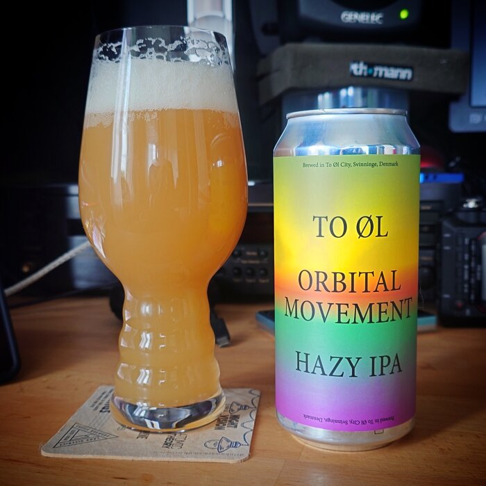 Ölglas med skummande hazy IPA och burk med etiketten "To Øl Orbital Movement Hazy IPA" på ett bord.