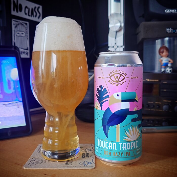 Glas med ljus, grumlig öl och en burk märkt "Toucan Tropic DDH Hazy IPA" på ett bord.