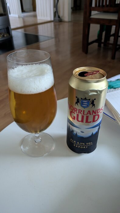 Ett glas öl bredvid en burk öl på ett bord inomhus.